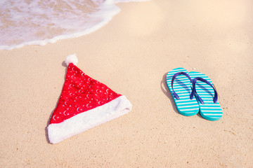 Fototapeta na wymiar Christmas holidays concept. Top view of flip flops and santa claus hat on sand beach.