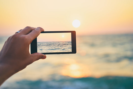 Cell technology and vacation. Close up of male hand holding smartphone, taking picture of sea sunset.