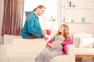 The husband holding cup of beverage for his pregnant wife while she is sitting on sofa at home. Happy pregnant family.