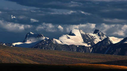 A highland meadows with yellow grass on a background of dramatic snow covered high mountains and glaciers under clouds and sunset sky, Plateau Ukok, Altai mountains, Siberia, Russia