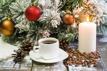 small white cup of coffee, roasted coffee beans, lighted candle, and fir branch in snow with cone, Christmas decorations on wooden background