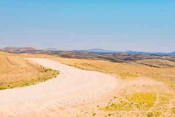 Fototapeta na wymiar Gravel winding road crossing the colorful Namib desert, in the majestic Namib Naukluft National Park, best travel destination in Namibia, Africa.