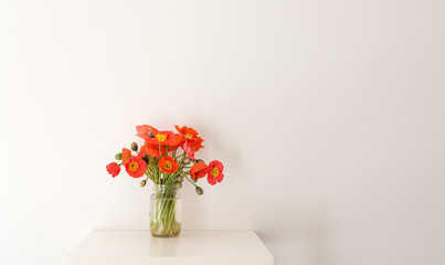 Red poppies in glass jar on white table against white wall