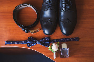 set of groom bow tie, boutonniere, belt, perfume and shoes lie on wooden table