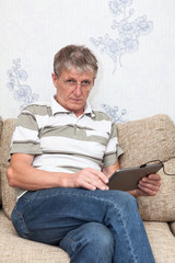 Caucasian senior man using tablet pad in domestic room sitting on sofa, looking at camera over glasses