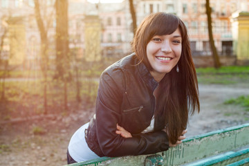 Smiling young woman walking on the spring park in sun rays, leaning to bench