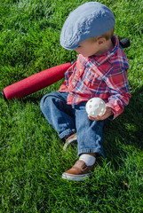 Toddler boy playing baseball with a white wiffle ball and red bat. Wearing blue motoring cap, red plaid shirt and blue jeans on grass on sunny cloudy day.