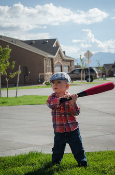 Toddler Boy Playing Baseball With A White Wiffle Ball And Red Bat. Wearing Blue Motoring Cap, Red Plaid Shirt And Blue Jeans On Grass On Sunny Cloudy Day.