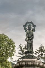 Fototapeta na wymiar Kyoto, Japan - September 16, 2016: Quan Yin standing in lotus and with halo statue on pedestal in Yuzen-en garden of Chion-in Buddhist Temple. Cloudy skies, some green trees.