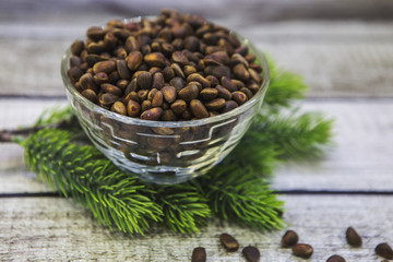 На деревянном столе лежит еловая ветка. На ветке стоит стеклянная ваза с кедровыми орехами. Рядом на столе россыпь орехов.