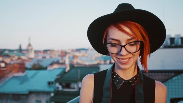 Young beautiful smiling girl taking picture on the street. Happy photographer on city background. Model wearing eyeglasses and stylish black wide-brimmed hat. Close up.