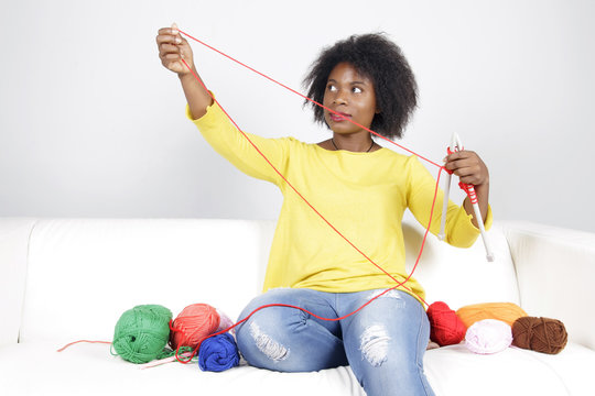 African woman knitting, sitting on a white couch. Surrounded by balls of yarn, she is knitting something with red colour.