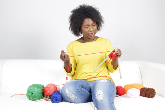 African woman knitting, sitting on a white couch. Surrounded by balls of yarn, she is knitting something with red colour.