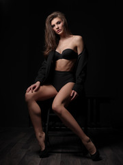 Fototapeta na wymiar Seductive, beautiful and attractive young female model with loose brown hair, sexy gorgeous figure and long legs in the black seamless underwear and wool coat is posing on the chair in the studio