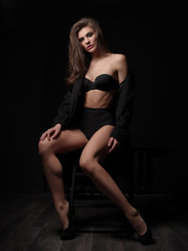 Seductive, beautiful and attractive young female model with loose brown hair, sexy gorgeous figure and long legs in the black seamless underwear and wool coat is posing on the chair in the studio