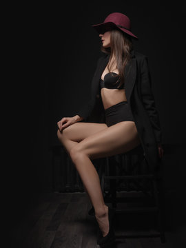 Appealing, elegant and attractive young female model with loose brown hair, sexy gorgeous figure and long legs in the black seamless underwear and wool coat is posing on the chair in the studio