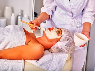 Woman middle-aged take facial and neck clay mask in spa salon. Interior with lot of cosmetic bottles.