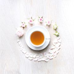 Upper sight of a cup of tea surrounded by cute small pink rose buds and its petals, white wood background. Closeup of cup of tea on wooden table. Flat lay.Still life with tea cup. Top View, copy-space