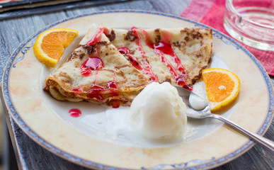 closeup shot of a dish plate with fritters pancakes, ice-cream ball, red topping, slices of orange and spoon