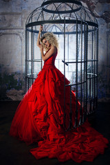 Fashion blonde in red dress with fluffy skirt near the birdcage, concept of liberation