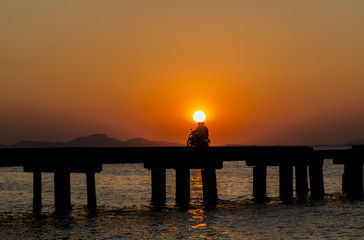 Big sun Sunset with silhouette people on motorcycle and long bridge on the beach in orange sky background