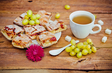 Homemade pie with jam on a plate, cup of tea, grapes, pieces of sugar and aster flower on wooden background. Snack or breakfast serving.