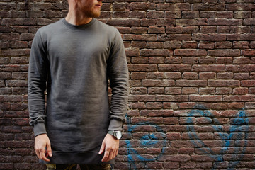 A cropped photo of a bearded hipster guy wearing blankl gray long sleeve t-shirt while standing on a brick wall background on a street. Empty place for you logo or design. - 123896254