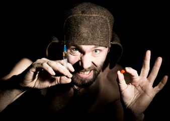 Dark portrait of scary evil sinister bearded man with smirk, offers a variety of drugs, a syringe or a pill. strange Russian man with a naked torso and a woolen hat