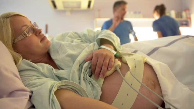 Two nurses are talking in the background while a pregnant woman is lying on the hospital bed peacefully. Close-up shot.
