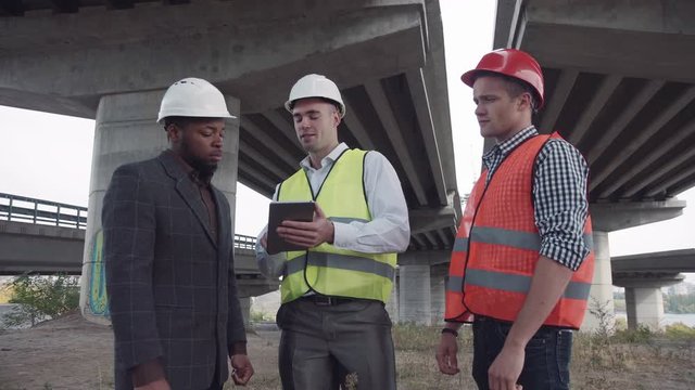 4K shot of structural engineers showing something on tablet to their bearded boss in white hard hat under newly constructed highway bridge