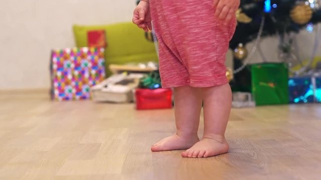 Little smiling baby boy feets run near garland on a background of Christmas tree and gift boxes with ribbon.
