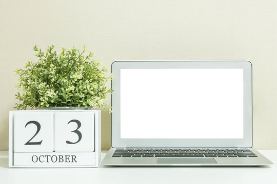 White wooden calendar with black 23 october word with white blank space at center of computer notebook on white wood desk and cream wallpaper textured background , selective focus at the calendar