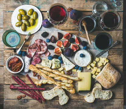 Wine and snack set with various wines in glasses, meat variety, bread, green olives, figs, nuts and berries on wax paper over rustic wooden background, top view, horizontal composition