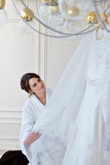 Beautiful bride in robe is watching a wedding dress. Beauty model girl in white clothes. Female portrait with bridal gown for marriage. Woman with curly hair and lace veil. Cute lady indoors