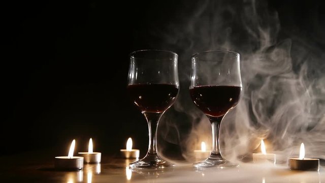 Two glasses of wine on black background and burning candles in the smoke.Red wine glasses.Glasses of red wine over candlelight and darkness.Romantic atmosphere with wine glasses and candles.Mystical