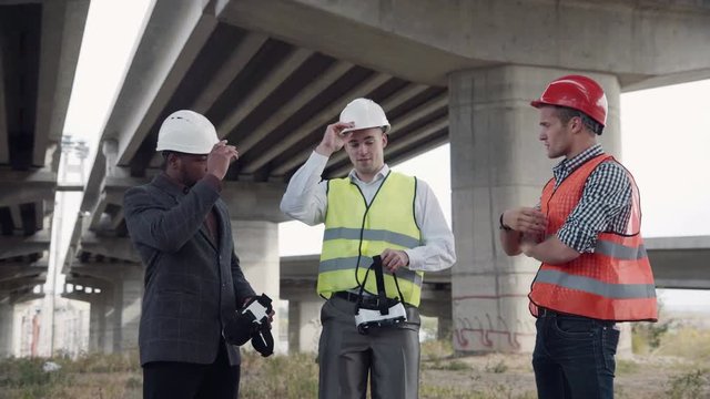 4K shot of business team of two architects and afro american foreman in suit uses virtual reality glasses to discuss a project of development while standing under overpass construction.