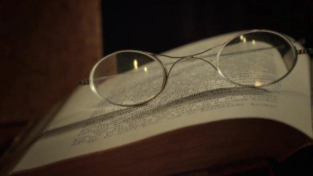 A pair of ancient glasses lying on an old book. Candles reflecting in glasses. Camera panning and moving.