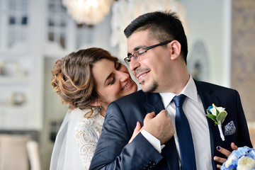 Wedding couple indoors is hugging each other. Beautiful model girl in white dress. Man in suit. Beauty bride with groom. Female and male portrait. Woman with lace veil. Cute lady and handsome guy