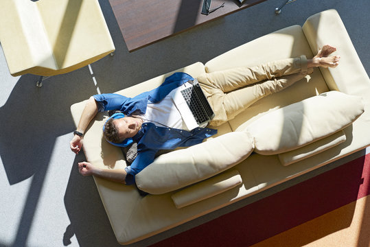 Top View Shot Of A Middle Aged Man With Laptop Lying On Couch And Listening Music While Relaxing At Home.