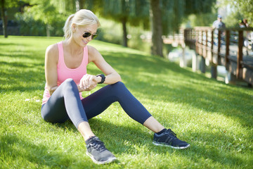 Naklejka na ściany i meble Running is my daily routine. Full length shot of a fit middle aged woman sitting in grass after morning workout and checking app on her sportwatch.
