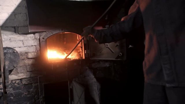 Man kindles fire in the furnace. The old boiler house. Maintenance of old steam boiler. stove oven. charcoal char. heat stoke hot fire. steam engine. nostalgic technology. smoke puffing funnel
