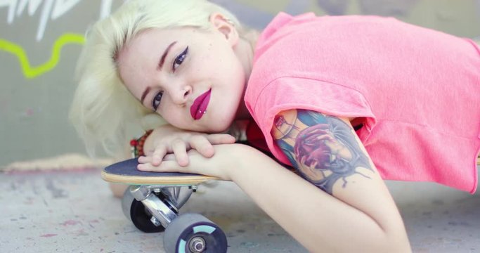 Pretty young blond woman with an arm tattoo lying resting her head on a skateboard looking at the camera  close up view