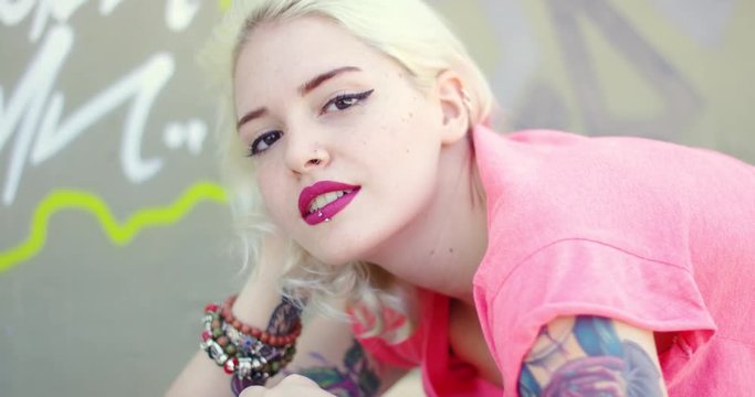 Gorgeous young blond woman with a vampire tattoo on her arm and a pierced lip lying relaxing on her skateboard at the skate park smiling at the camera.