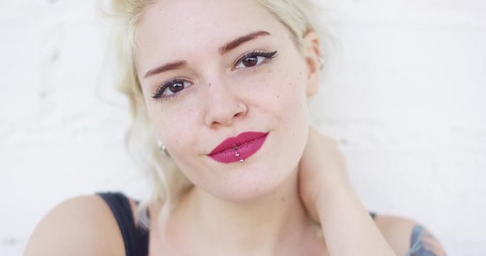 Attractive blond woman with a lip piercing wearing red lipstick smiling quietly at the camera  close up frontal head and shoulders