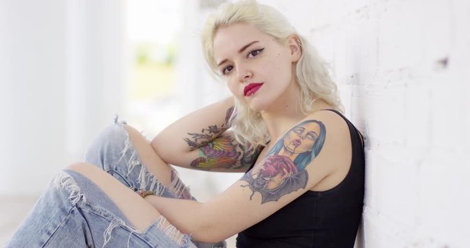Sexy sultry young woman with a vampire tattoo on her arm sitting leaning against a white outdoor wall turning to stare at the camera