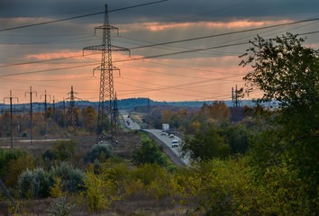 Road with High voltage power pylons. On the road driving cars. Came the autumn, the leaves turn yellow in the trees, and the rain clouds on the sky