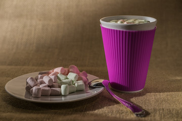 vivid pink colored cup with sweet drink and marshmallows on top near white plate with bunch from small and different colored marshmallows with spoon on foreground and burlap bacground/melt