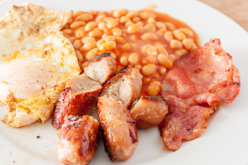 A fried English breakfast of sausges, bacon, fried and baked beams, on a white plate