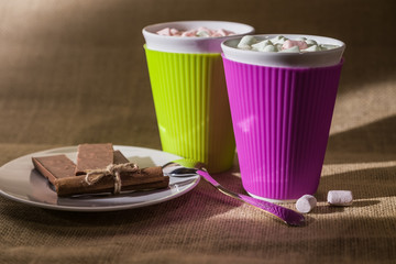 couple of vivid colored cups with sweet drink and marshmallows on top near white round plate with chocolate and cinnamon roll and spoon all on burlap background