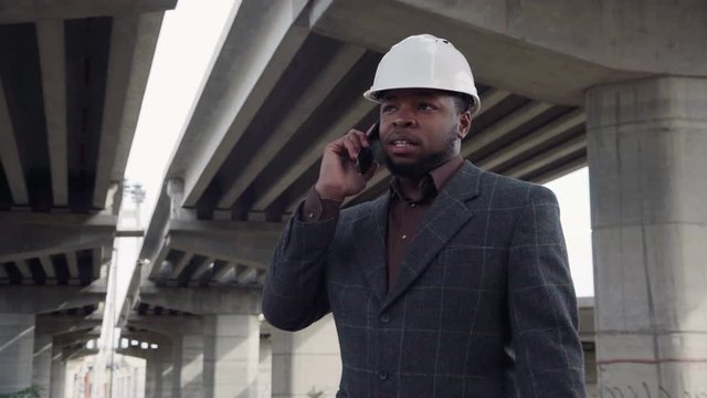 Smiling african man wearing white protective helmet talking on mobile phone while standing under the overpass construction and then looking at camera